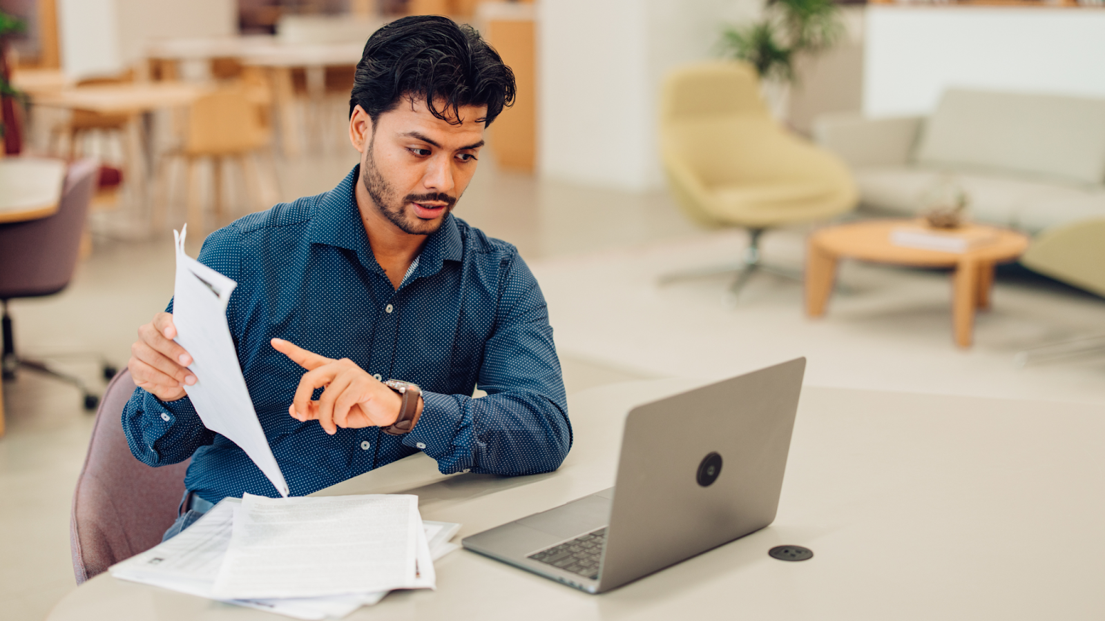 A transaction coordinator working at a desk, holding documents and pointing at them while looking at a laptop, suggesting focus and coordination in a modern office environment.
